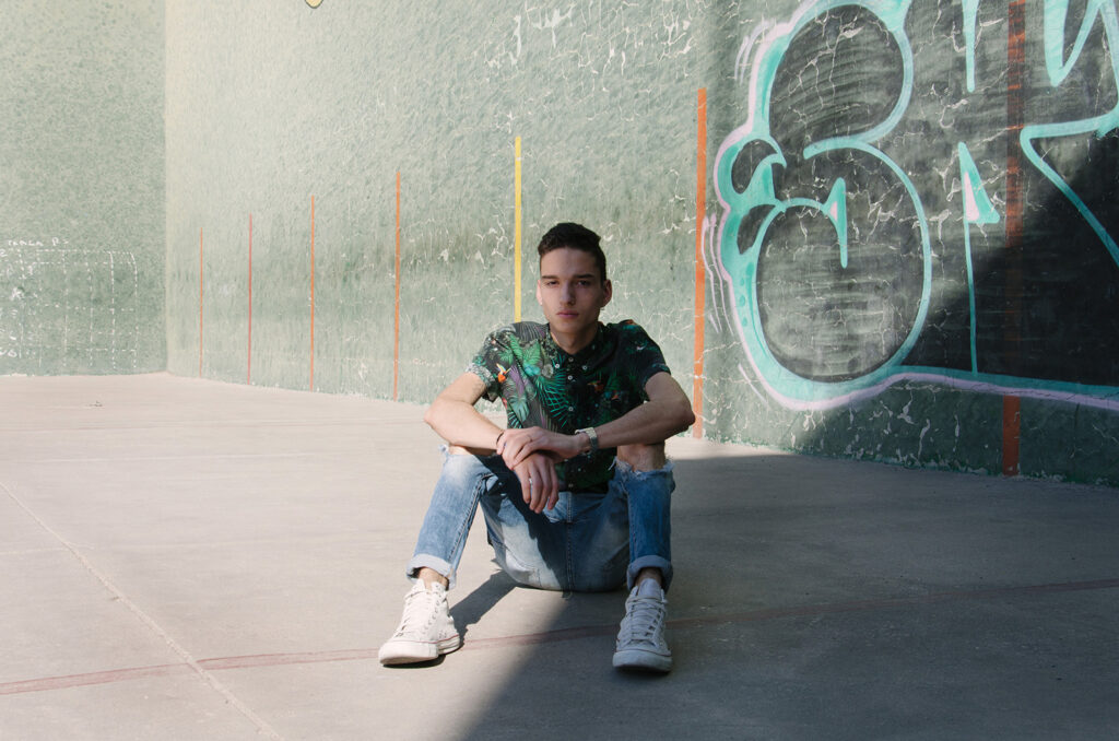Victor Zepeda sits against a graffiti-covered wall wearing a tropical shirt and blue jeans with a watch on his wrist.
